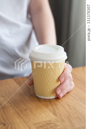 Woman holding paper coffee cup with white lid on wooden table in natural daylight. 131378307