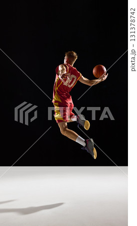 Basketball player in red jersey soaring in jump holding ball with determination in dark background Basketball player in red jersey soaring in jump holding ball with determination in dark background 131378742