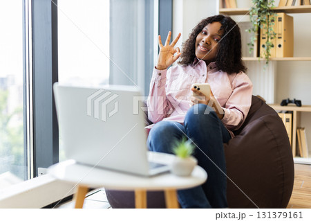 Young adult woman sitting on beanbag holding smartphone gesturing OK sign. Cozy home office environment with laptop, bright window, bookshelf. Concept of confidence work-life balance positivity 131379161