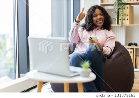 Young African woman in her twenties sitting indoors using smartphone, smiling and gesturing approval. Modern workspace with natural light relaxed, productive, and positive atmosphere. Young African woman in her twenties sitting indoors using smartphone, smiling and gesturing approval. Modern workspace with natural light relaxed, productive, and positive atmosphere. 131379163