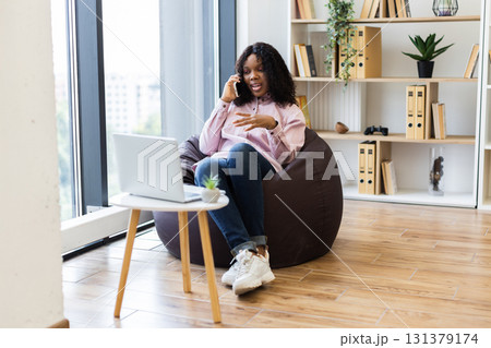 Smiling young woman sitting on bean bag using smartphone and laptop in cozy office. Relaxed atmosphere with shelves, plants, and natural light. Female gracefully balancing work and communication. Smiling young woman sitting on bean bag using smartphone and laptop in cozy office. Relaxed atmosphere with shelves, plants, and natural light. Female gracefully balancing work and communication. 131379174