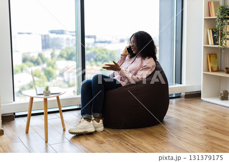 Young adult woman sitting on brown beanbag near window, talking on smartphone, casual ambiance. Modern living space includes wooden floors, small table with laptop and plant, emphasizing comfort. Young adult woman sitting on brown beanbag near window, talking on smartphone, casual ambiance. Modern living space includes wooden floors, small table with laptop and plant, emphasizing comfort. 131379175