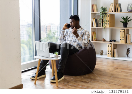 Young adult male sitting on beanbag chair in modern home office multitasking with laptop and phone Young adult male sitting on beanbag chair in modern home office multitasking with laptop and phone 131379181