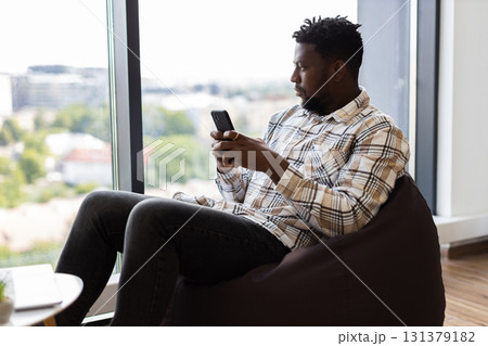 Young adult male sitting on brown beanbag, using smartphone and laptop, modern leisure concept in bright indoor setting. Young adult male sitting on brown beanbag, using smartphone and laptop, modern leisure concept in bright indoor setting. 131379182