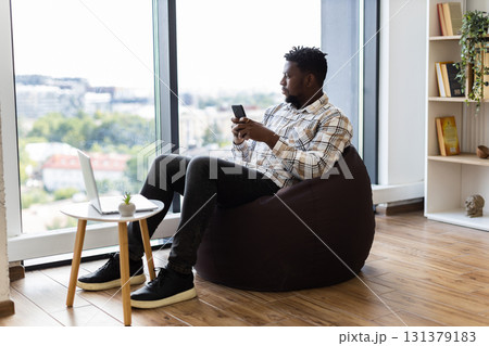 Young adult male sitting on brown beanbag, using smartphone and laptop, modern leisure concept in bright indoor setting. Young adult male sitting on brown beanbag, using smartphone and laptop, modern leisure concept in bright indoor setting. 131379183