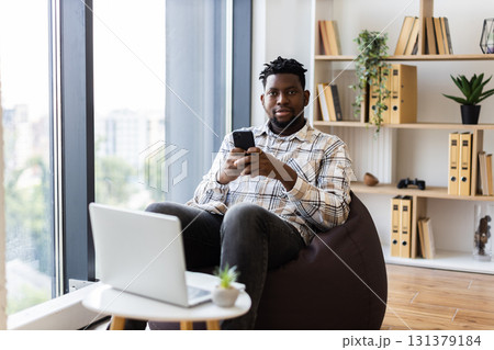 Young male adult using smartphone while sitting on beanbag, laptop on table nearby, indoors 131379184