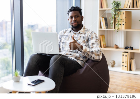 Young adult man sitting on beanbag in modern home office, working on laptop, showing positive gesture. Bright setting with natural light and organized shelves in background creating calm workspace. 131379196