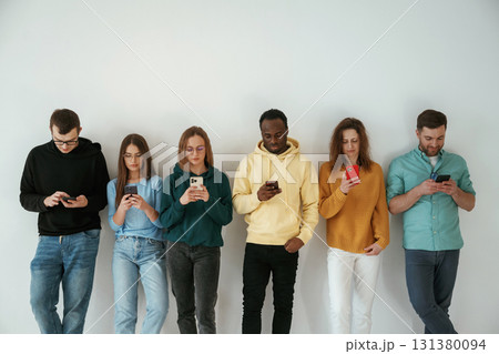 Busy by using smartphones. Group of young people are standing against white background Busy by using smartphones. Group of young people are standing against white background 131380094