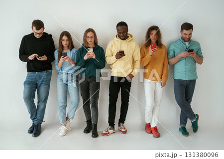 Busy by using smartphones. Group of young people are standing against white background Busy by using smartphones. Group of young people are standing against white background 131380096
