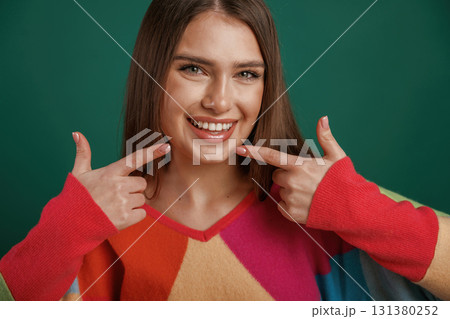 Pointing to the teeth by fingers, smiling. Young woman is standing against green background in the studio Pointing to the teeth by fingers, smiling. Young woman is standing against green background in the studio 131380252