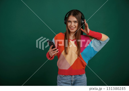 Moment of enjoying music, in headphones. Young woman is standing against green background in the studio 131380285