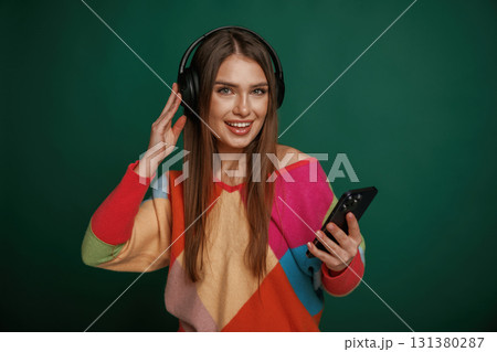 Moment of enjoying music, in headphones. Young woman is standing against green background in the studio Moment of enjoying music, in headphones. Young woman is standing against green background in the studio 131380287