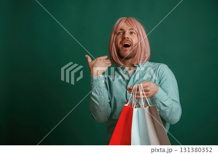 Posing, hand near face. Holding shopping bags. Gay man with pink hair is standing against green background Posing, hand near face. Holding shopping bags. Gay man with pink hair is standing against green background 131380352