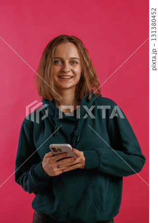 With smartphone. Young woman is standing against red background in the studio With smartphone. Young woman is standing against red background in the studio 131380452
