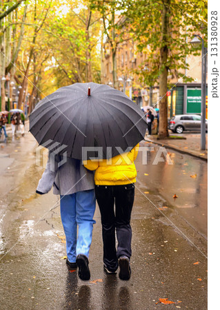 Two people walk together under a large umbrella on a rainy city street with autumn trees and wet pavement. Concept of umbrella protection, togetherness, rainy weather, autumn season 131380928