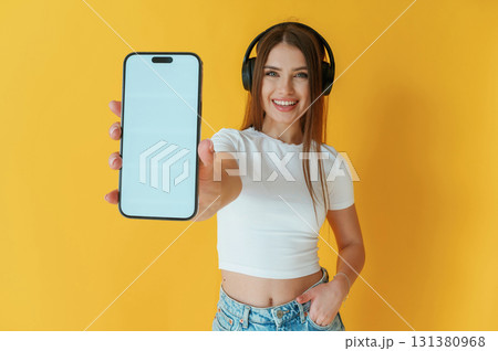 Headphones on head, holding smartphone with copy space. Young woman is against yellow background Headphones on head, holding smartphone with copy space. Young woman is against yellow background 131380968