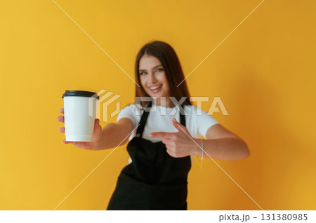 Black and white coffee worker uniform, holding cup of drink. Young woman is against yellow background Black and white coffee worker uniform, holding cup of drink. Young woman is against yellow background 131380985