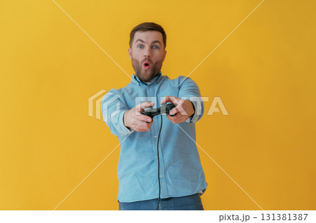 Playing video game with joystick. Attractive man in blue shirt is against yellow background 131381387