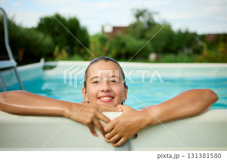 Teen girl enjoys summer fun while peeking over the edge of a swimming pool in a sunny backyard setting Teen girl enjoys summer fun while peeking over the edge of a swimming pool in a sunny backyard setting 131381580