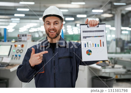 Plan with graphs in hand. Factory worker is indoors with hard hat 131381798