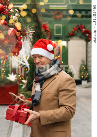 Adult happy man in Santa hat buying red gift at Christmas outdoor market. Xmas shopping. Side view. Holiday spirit. Close up. Adult happy man in Santa hat buying red gift at Christmas outdoor market. Xmas shopping. Side view. Holiday spirit. Close up. 131383469