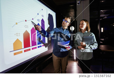 Conception of profit. Man and woman are standing near projector with business data, cooperation Conception of profit. Man and woman are standing near projector with business data, cooperation 131387102