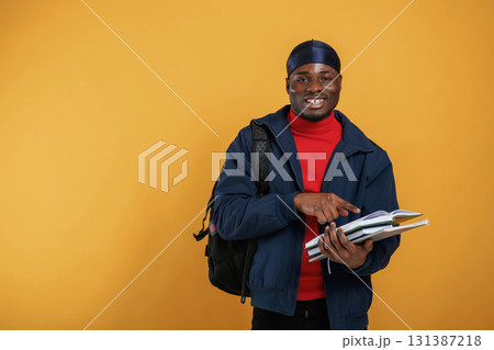 Student with books, backpack and notepad. Handsome black man is in the studio against yellow background 131387218