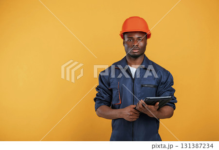 Digital tablet in hand, construction worker. Handsome black man is in the studio against yellow background 131387234