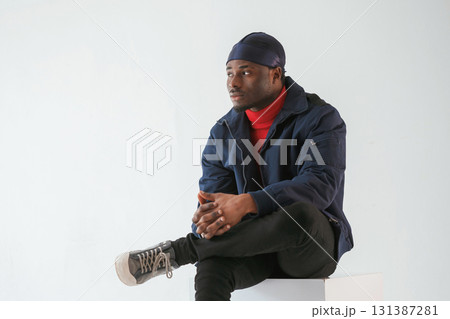 Sitting on the cube chair. Black man in cap is in the studio against white background 131387281