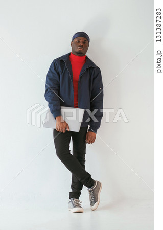 Standing and holding laptop in hand. Black man in cap is in the studio against white background 131387283