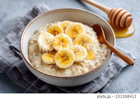 A bowl of oatmeal topped with sliced bananas and drizzled with honey served with a wooden spoon. 131388411