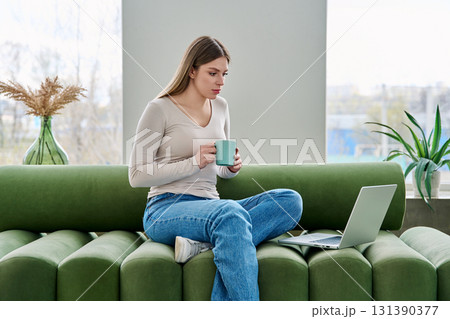 Young relaxing woman with cup looking at laptop screen, sitting on sofa at home 131390377
