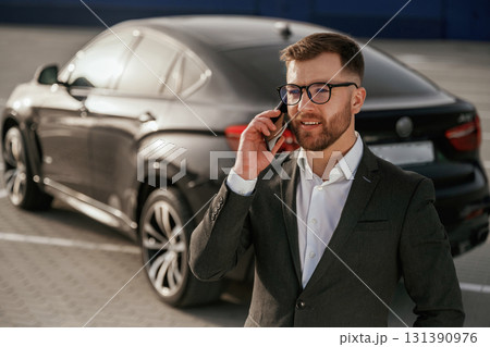 Talking by using phone. Businessman in suit is near his black car outdoors Talking by using phone. Businessman in suit is near his black car outdoors 131390976