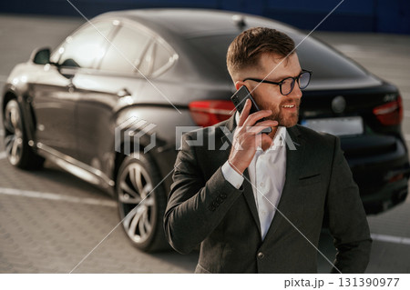 Cheerful facial expression. Businessman in suit is near his black car outdoors Cheerful facial expression. Businessman in suit is near his black car outdoors 131390977
