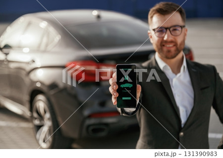 Charging process icon in the smartphone. Businessman in suit is near his black car outdoors Charging process icon in the smartphone. Businessman in suit is near his black car outdoors 131390983