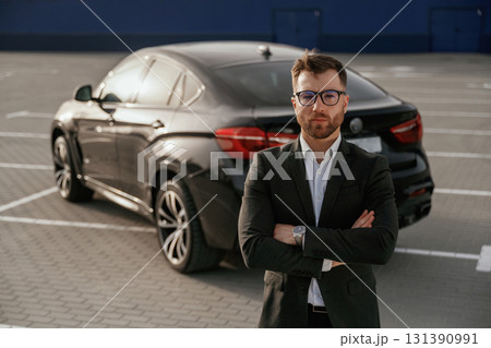 Standing with arms crossed. Businessman in suit is near his black car outdoors 131390991