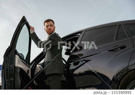 Natural lighting, day. Businessman in suit is near his black car outdoors 131390995