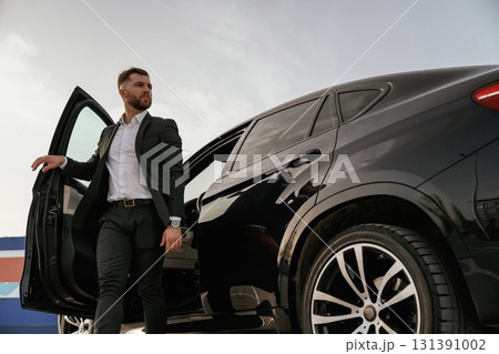 Natural lighting, day. Businessman in suit is near his black car outdoors 131391002