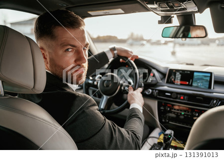 Looking behind at the camera. Man in black suit is sitting in the car, driving Looking behind at the camera. Man in black suit is sitting in the car, driving 131391013