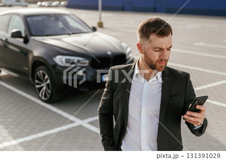 Smartphone in hand. Businessman in suit is near his black car outdoors Smartphone in hand. Businessman in suit is near his black car outdoors 131391029