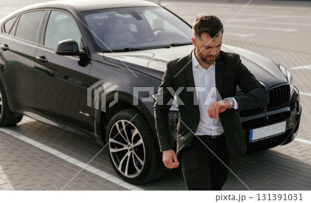 Checks the time. Businessman in suit is near his black car outdoors Checks the time. Businessman in suit is near his black car outdoors 131391031