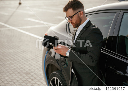 Leaning on the vehicle while it charges, with smartphone. Businessman in suit is near his black car outdoors Leaning on the vehicle while it charges, with smartphone. Businessman in suit is near his black car outdoors 131391057