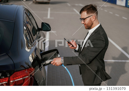 Charging the electric vehicle. Businessman in suit is near his black car outdoors 131391070