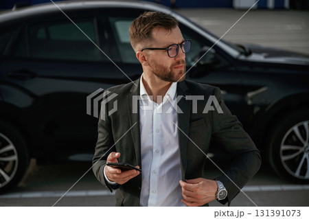 Smartphone in hand. Businessman in suit is near his black car outdoors Smartphone in hand. Businessman in suit is near his black car outdoors 131391073