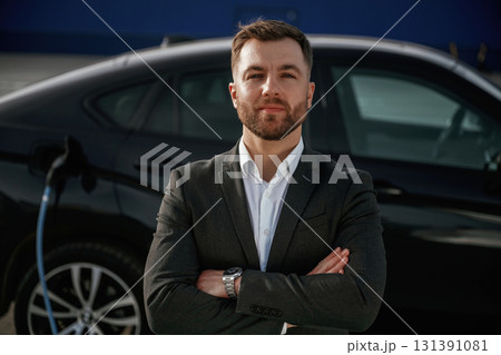 Standing with arms crossed. Businessman in suit is near his black car outdoors Standing with arms crossed. Businessman in suit is near his black car outdoors 131391081