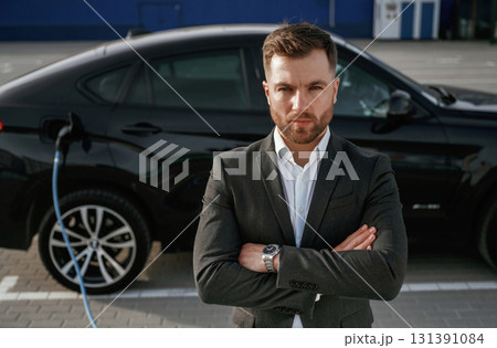 Front view, arms crossed. Businessman in suit is near his black car outdoors Front view, arms crossed. Businessman in suit is near his black car outdoors 131391084