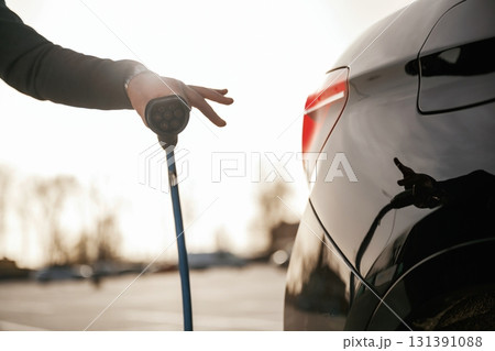 Blue wire for charging is in hand. Businessman in suit is near his black car outdoors 131391088