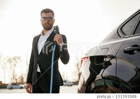 Blue wire for charging is in hand. Businessman in suit is near his black car outdoors 131391089