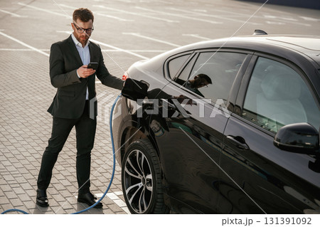 Blue wire for charging is in hand. Businessman in suit is near his black car outdoors Blue wire for charging is in hand. Businessman in suit is near his black car outdoors 131391092