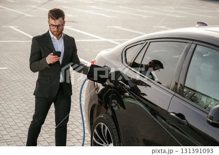 Holding phone in hand while vehicle is charging. Businessman in suit is near his black car outdoors 131391093